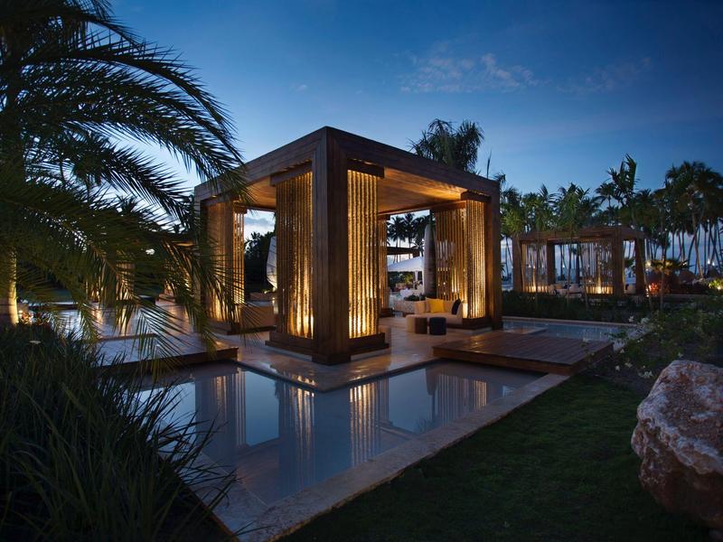 Ein beleuchteter moderner Pavillon mit Wasserbecken umgeben von Palmen und Garten bei Dämmerung.