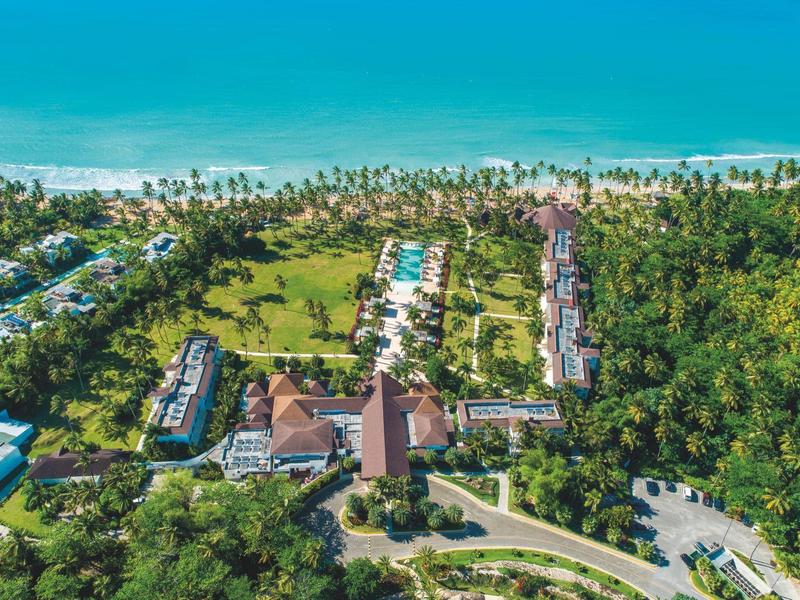 Luftaufnahme eines Resorts mit Pool, umgeben von Palmen und dem blauen Meer im Hintergrund.