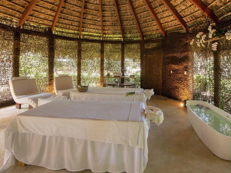 Innenraum einer Massagehütte mit Strohdach, zwei Massagetischen, weißem Wannne und Stühlen.