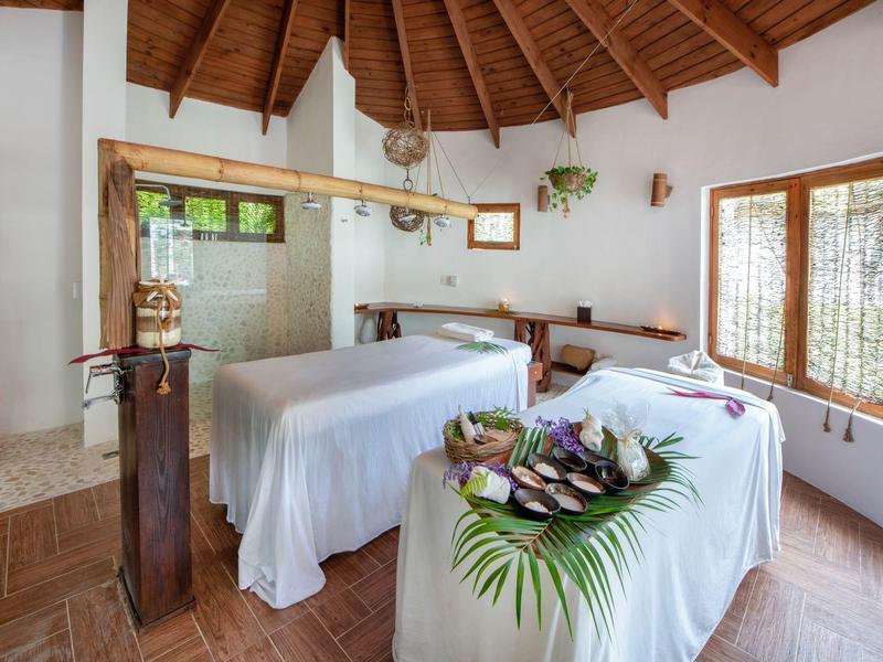 Innenraum eines hellen Massageraums mit Holzdach, zwei Massageliegen und Dekoration aus Pflanzen.