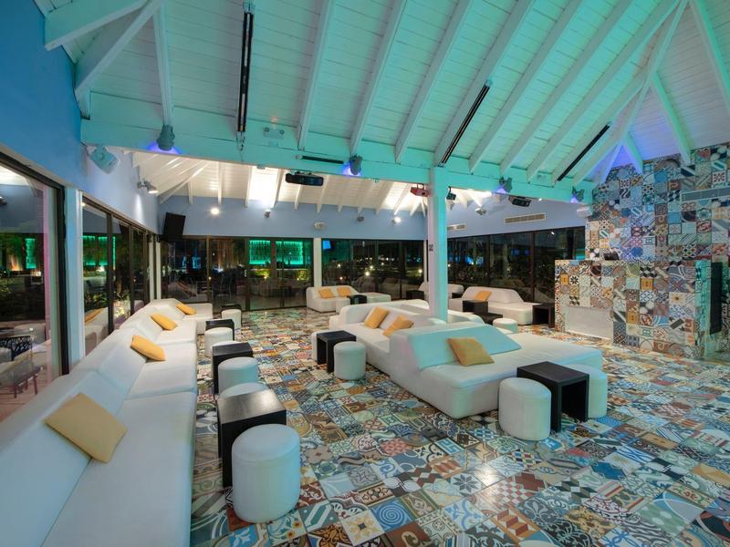 Offener, moderner Lounge-Bereich mit bunten Fliesen, weißen Sitzmöbeln und einem bedachten Dach.