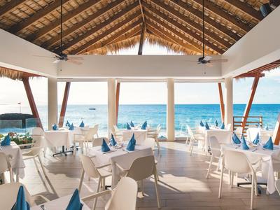 Offenes Restaurant mit Blick aufs Meer, Tischen mit weißen Stühlen und blauen Servietten unter einem Holzdach.