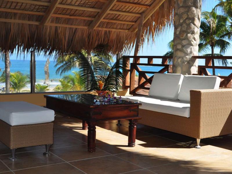 Outdoor-Lounge mit Sofa, Hocker und Tisch unter Strohdach, Palmen und Meer im Hintergrund.