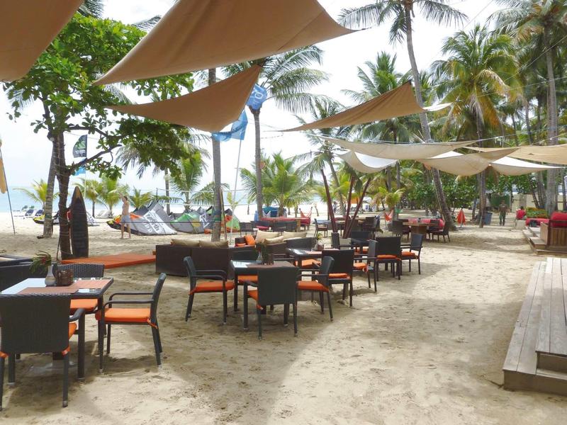 Strandrestaurant mit Tischen, Stühlen und Sonnensegeln unter Palmen am Sandstrand.