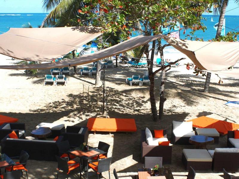 Strand mit weißem Sand, Palmen und Sonnensegeln über Tischen und Sitzgruppen in Schwarz und Orange.