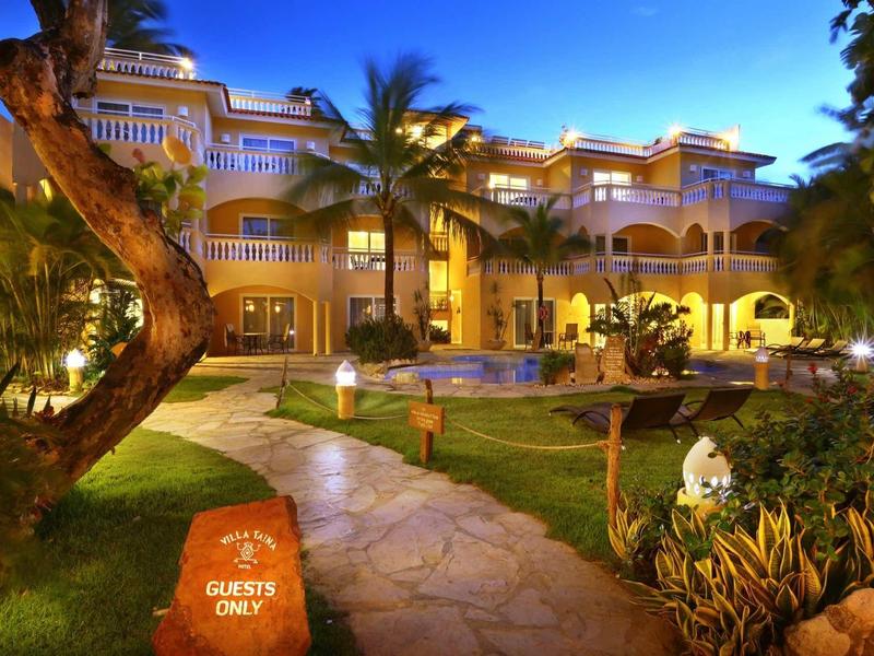 Großes beleuchtetes Hotel mit Palmen, Gehweg, Garten und Schwimmbad am Abend unter blauem Himmel.