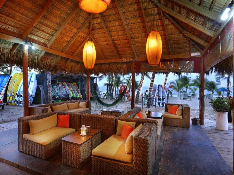 Offener Lounge-Bereich mit Holzdach, gemütlichen Sitzmöbeln und orangefarbenen Lampen am Strand.