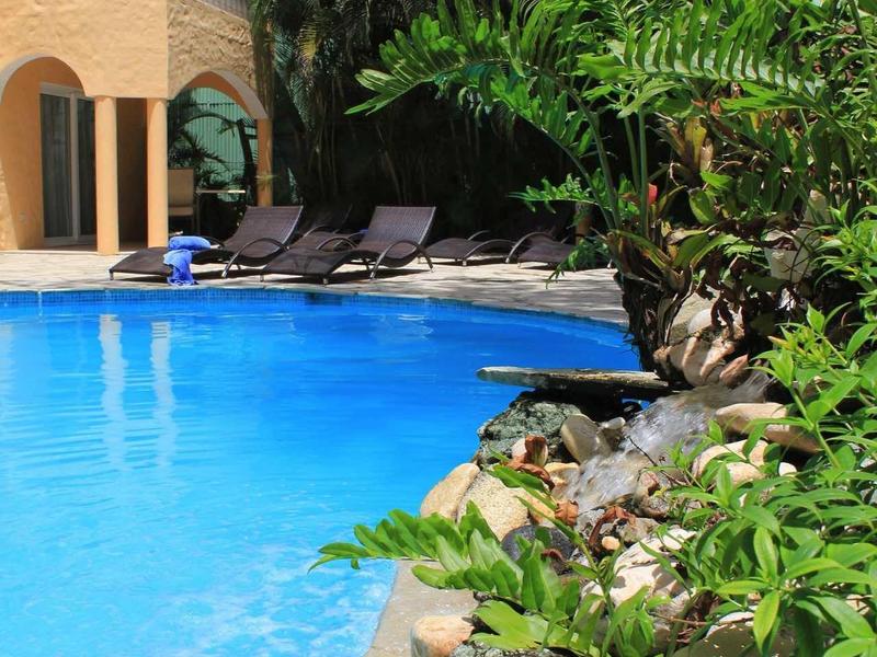 Blauer Pool neben Palmen und grünen Pflanzen vor einem gelben, mediterranen Haus mit Balkon.