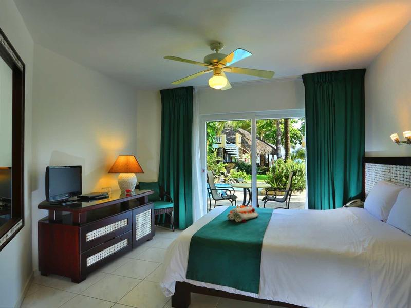 Heller Hotelzimmer mit großem Bett, grünem Dekor, Ventilator und Blick auf einen Pool durch Fenster.