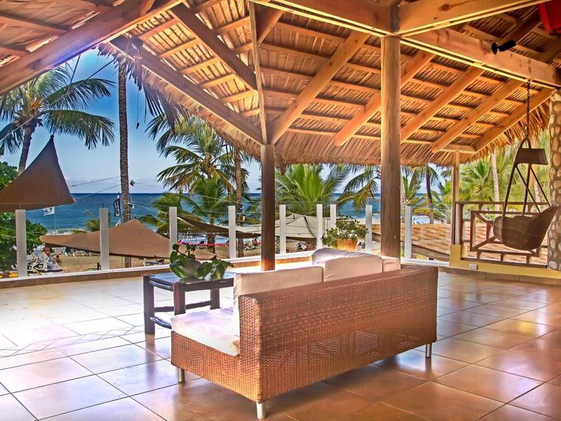 Offener Pavillon mit Holzdach, hellen Fliesen, Pflanzen und Blick auf Palmen und Meer.