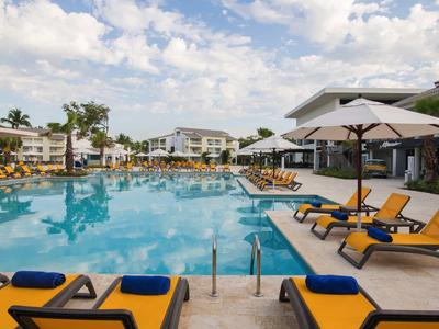 Groot hotelzwembad met ligstoelen onder parasols en moderne gebouwen op de achtergrond.