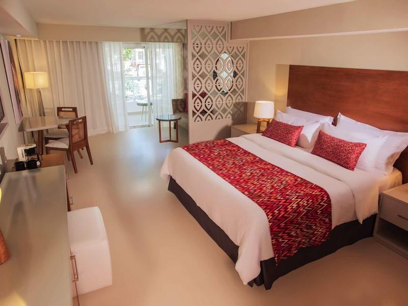 Geräumiges Hotelzimmer mit großem Bett, roten Akzenten, Schreibtisch und Balkon mit Vorhängen.