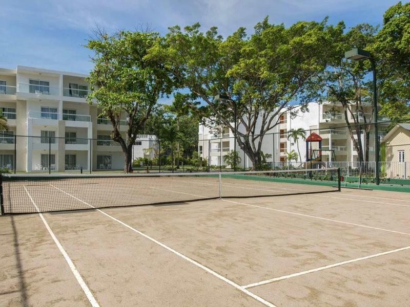 Leerer Sand-Tennisplatz mit Netz, umgeben von Bäumen und mehrstöckigen weißen Gebäuden.