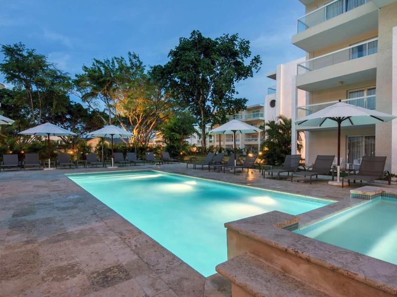 Beleuchteter Pool mit Liegestühlen und Sonnenschirmen neben modernem Gebäude bei Abenddämmerung.