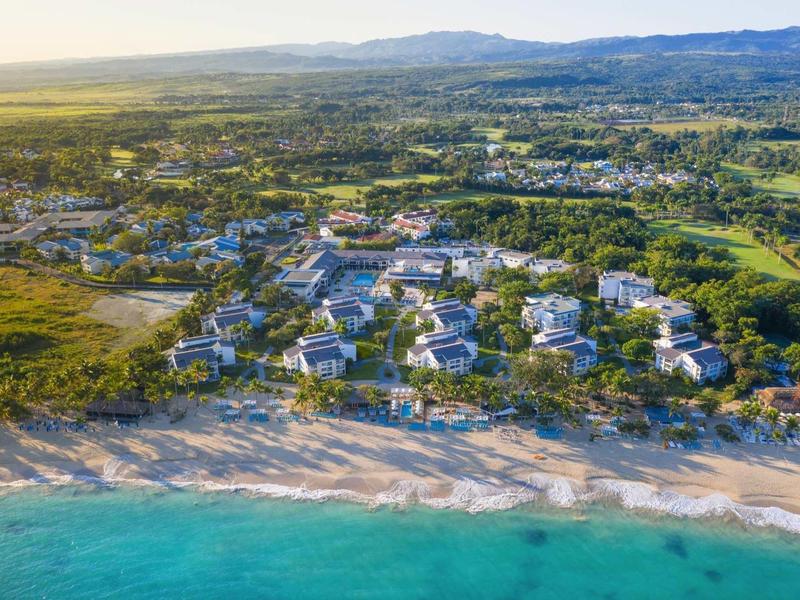 Luftaufnahme einer Küstenstadt mit Strand, blauem Meer, weißen Gebäuden und grüner Landschaft dahinter.