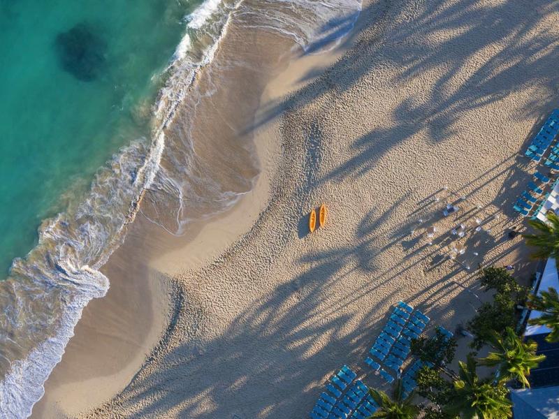 Vogelperspektive auf einen Strand mit türkisblauem Wasser, Sand, Schatten und Strandliegen.