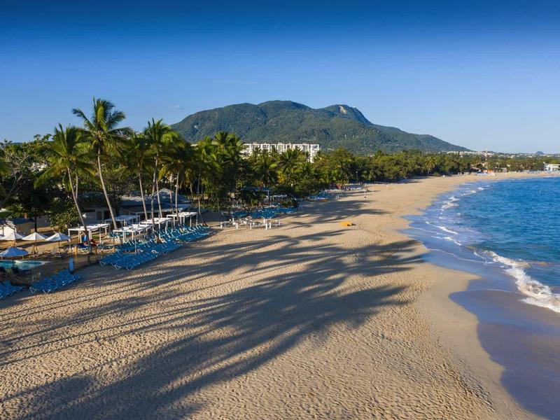 Weitläufiger Sandstrand mit Palmen, klarem blauem Meer und Bergen im Hintergrund bei sonnigem Himmel.