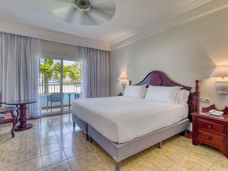 Helles Hotelzimmer mit großem Bett, Ventilator an der Decke und Blick auf Balkon mit Palmen.