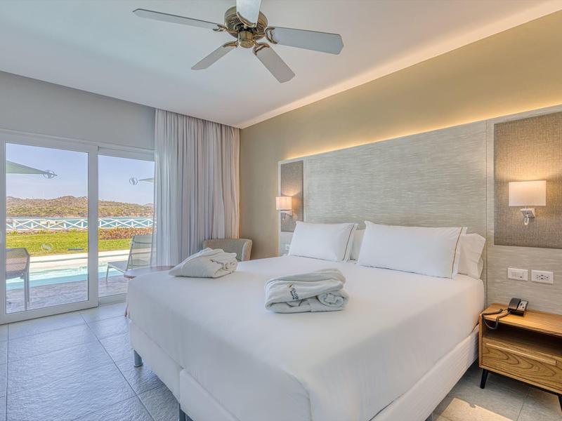 Helles Hotelzimmer mit Doppelbett, Deckenventilator und Balkon mit Blick auf die Landschaft.