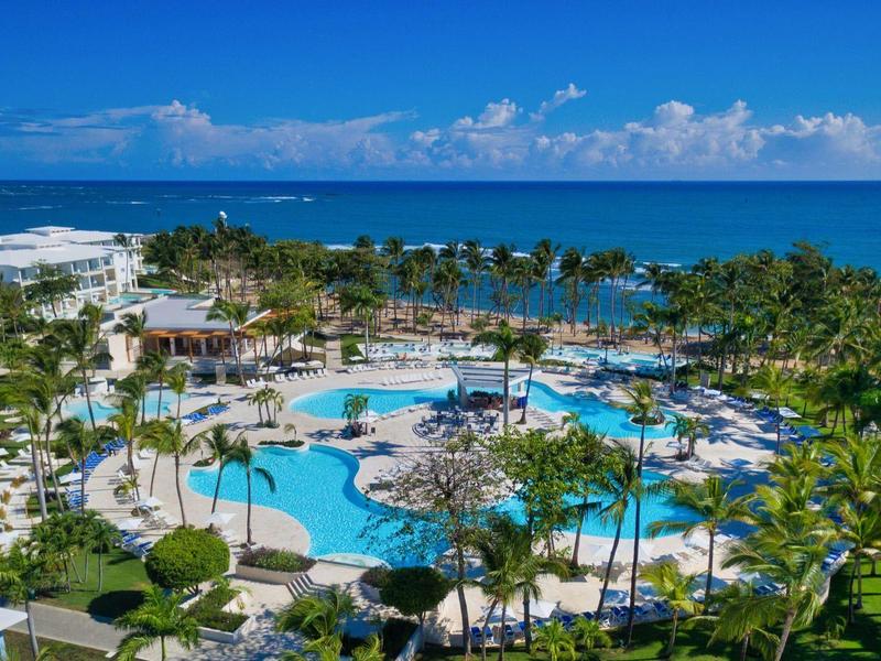 Große Poolanlage mit Palmen, Liegestühlen und Blick auf das blaue Meer unter klarem Himmel.
