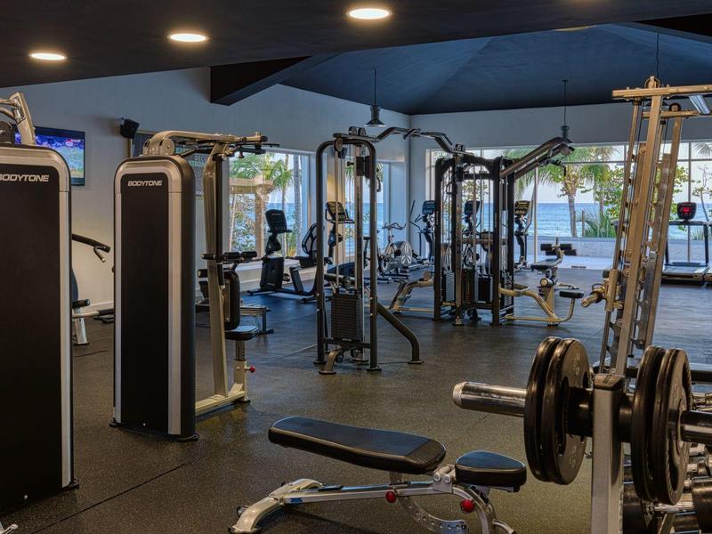 Moderner Fitnessraum mit verschiedenen Trainingsgeräten, Hanteln und großen Fenstern für Tageslicht.