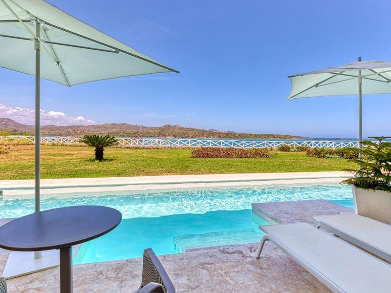 Moderner Außenbereich mit Pool, weißen Liegen, zwei Sonnenschirmen und Blick auf Berglandschaft.