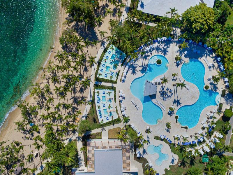 Luftaufnahme von Resort mit geschwungenem Pool, Liegen und grünem Strand unter klarem Himmel.