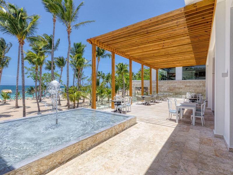 Moderner Balkon mit Holzüberdachung, Sitzmöbeln und Blick auf Pool, Palmen und Meer im Hintergrund.