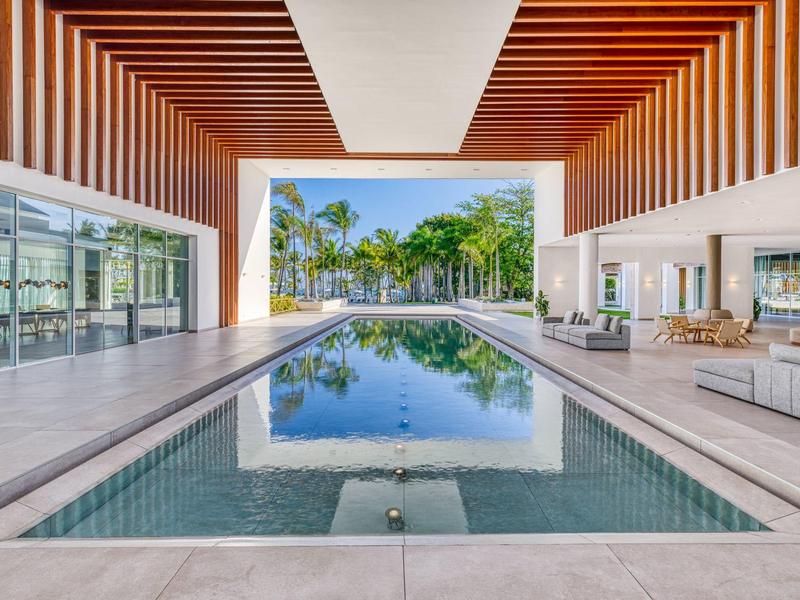 Moderner Pool mit Holzdecke, Glasfronten und Blick auf Palmen und blauen Himmel im Hintergrund.