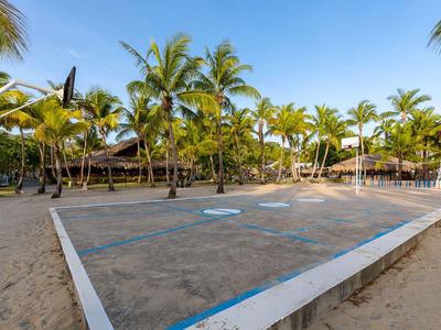 Sandige Sportanlage mit Basketballkorb und Volleyballnetz vor Palmen unter blauem Himmel.