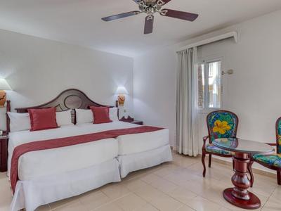 Helles Schlafzimmer mit großem Bett, Nachttischen, Deckenventilator und zwei farbigen Stühlen am Tisch.