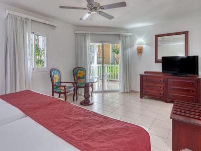 Helles Schlafzimmer mit weißem Bett, roter Bettdecke, zwei Stühlen, Kommode und Balkonzugang.