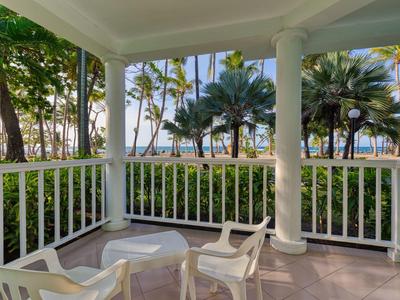 Überdachte Terrasse mit weißen Stühlen und Tisch, umgeben von Palmen und Blick aufs Meer.