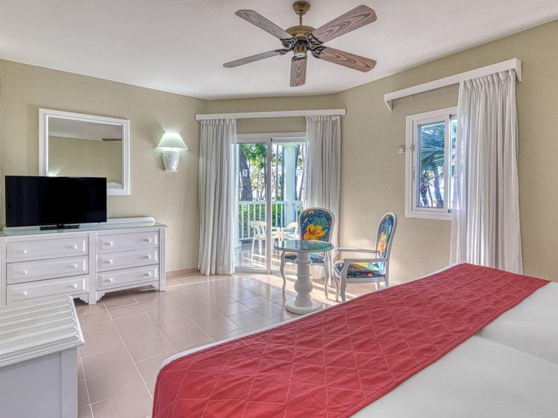 Helles Schlafzimmer mit Deckenventilator, rote Bettdecke, weißen Möbeln und Balkon mit Blick ins Grüne.