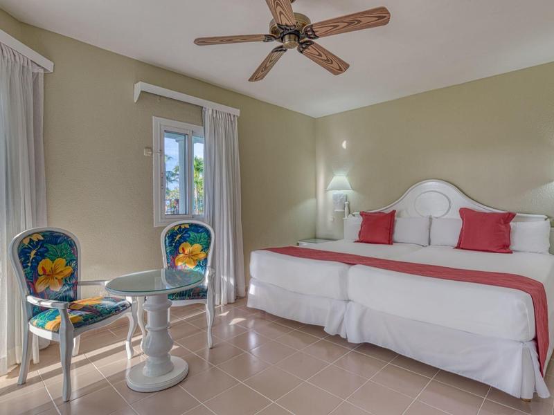 Helles Schlafzimmer mit großem Bett, weißen Möbeln, zwei bunten Stühlen und Deckenventilator.