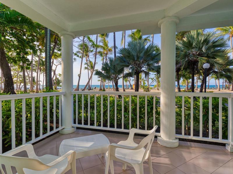 Überdachte Terrasse mit weißen Stühlen und Tisch, umgeben von Palmen und Blick aufs Meer.