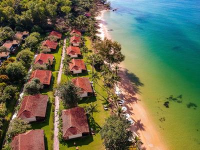 Luftaufnahme von Strandbungalows mit roten Dächern neben klarem, grünlichem Wasser und Sandstrand.