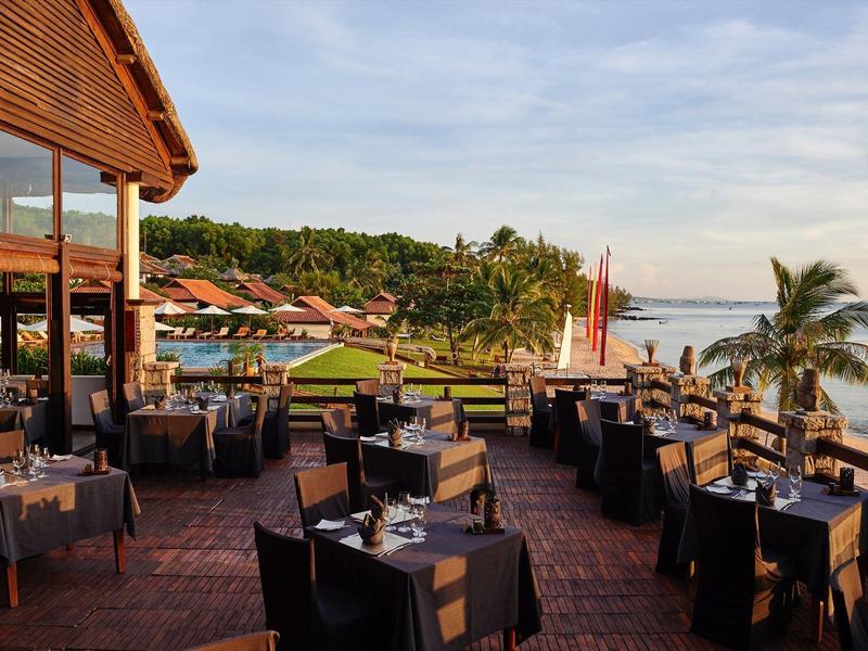 Strandrestaurant mit gedeckten Tischen, schwarzer Stühle, Holzdach und Blick auf Meer und Palmen.