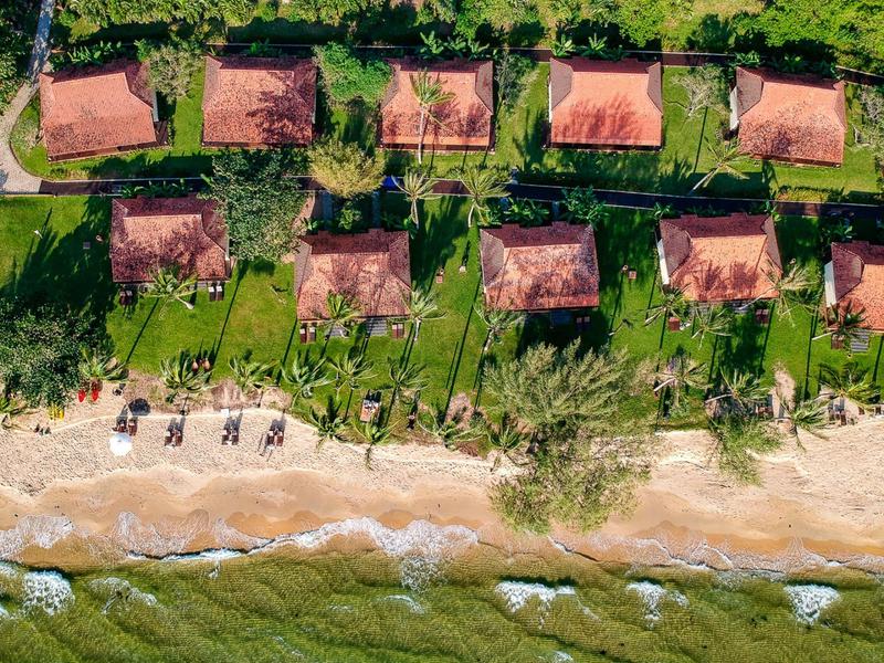 Rote Dächer, grüne Gärten und Palmen entlang eines sandigen Strandes mit blauem Meer.
