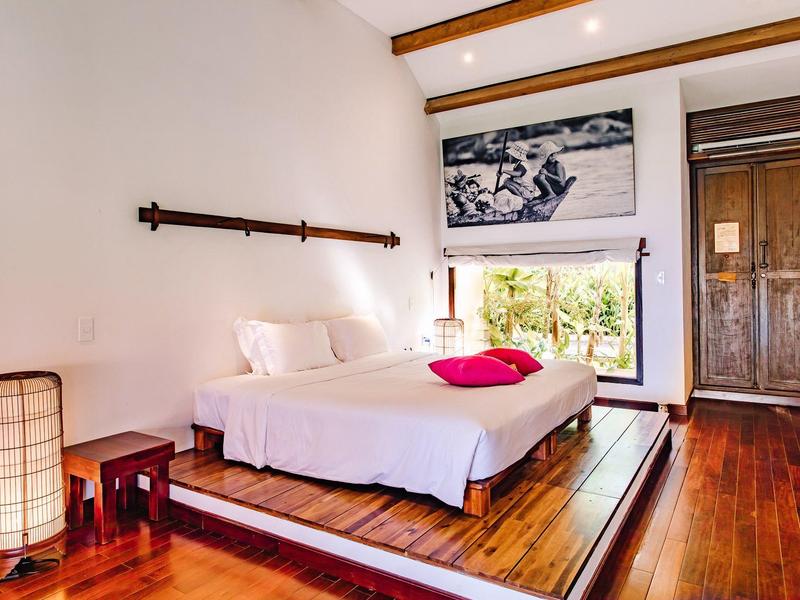 Helles Schlafzimmer mit Holzfußboden, Doppelbett, roter Decke, Holzschrank und großem Fenster.