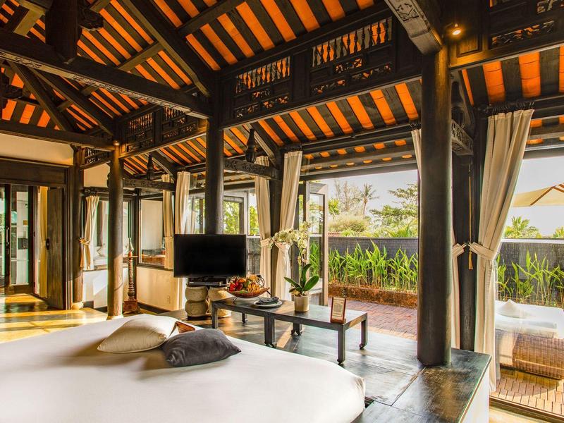 Offenes Schlafzimmer mit Holzbalken, großem Bett, TV und Blick auf grüne Pflanzen im Freien.