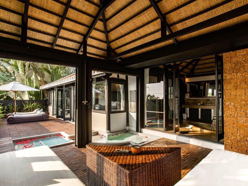 Offener Wohnbereich mit Strohdach, braunen Möbeln und Blick auf Terrasse mit Pflanzen und Sonnenliegen.