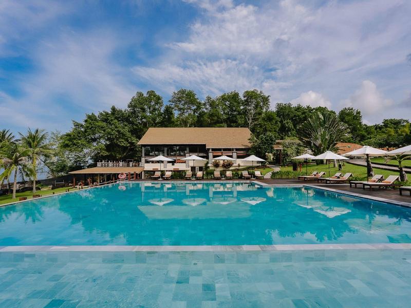 Großer, blauer Pool mit Liegestühlen, Sonnenschirmen und Palmendekor unter blauem Himmel.