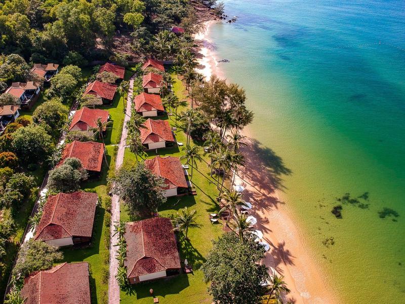 Reihen von kleinen Häusern mit roten Dächern am grünen Strand neben blauem Wasser und Bäumen.