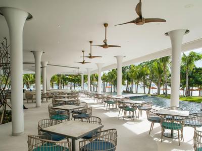 Offener moderner Außenbereich mit Tischen und Stühlen unter einer überdachten Terrasse, umgeben von Bäumen.