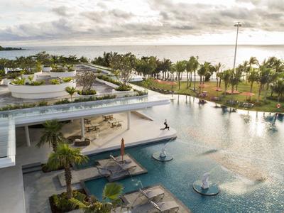 Área de piscina moderna con tumbonas y vista al mar y palmeras bajo cielo nublado.