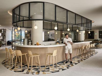 Área de bar moderna de hotel con taburetes de madera y un camarero con uniforme blanco.