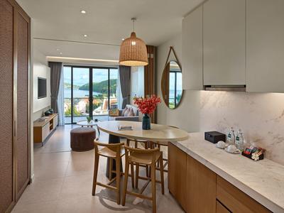 Área moderna de habitación de hotel con cocina, mesa comedor, sofá y vista al mar a través de grandes ventanas.