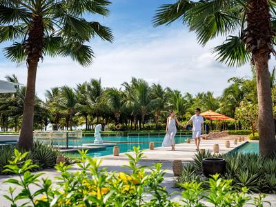 Poolbereich mit Palmen und Gästen, die auf dem Hotelgelände bei sonnigem Himmel spazieren