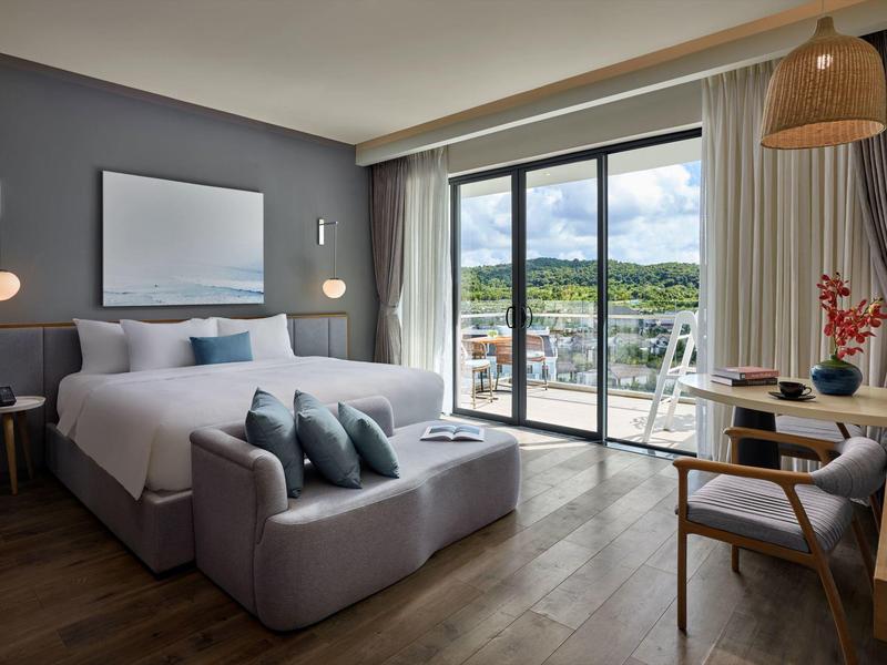 Habitación de hotel moderna con cama grande, balcón y vista a paisaje verde bajo cielo nublado.
